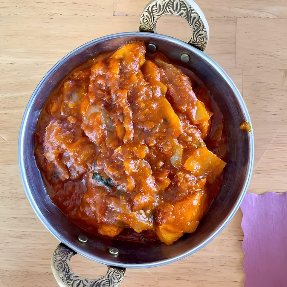Indian chicken curry in a metal bowl with ornate handles on a wooden table.
